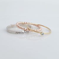 The set includes a silver ring with a brushed texture and engraved in cursive, a delicate rose gold beaded ring, and a thin yellow gold ring with a small bezel-set blue gemstone. The rings are arranged in an overlapping manner, creating a visually appealing composition with soft reflections on the surface below.