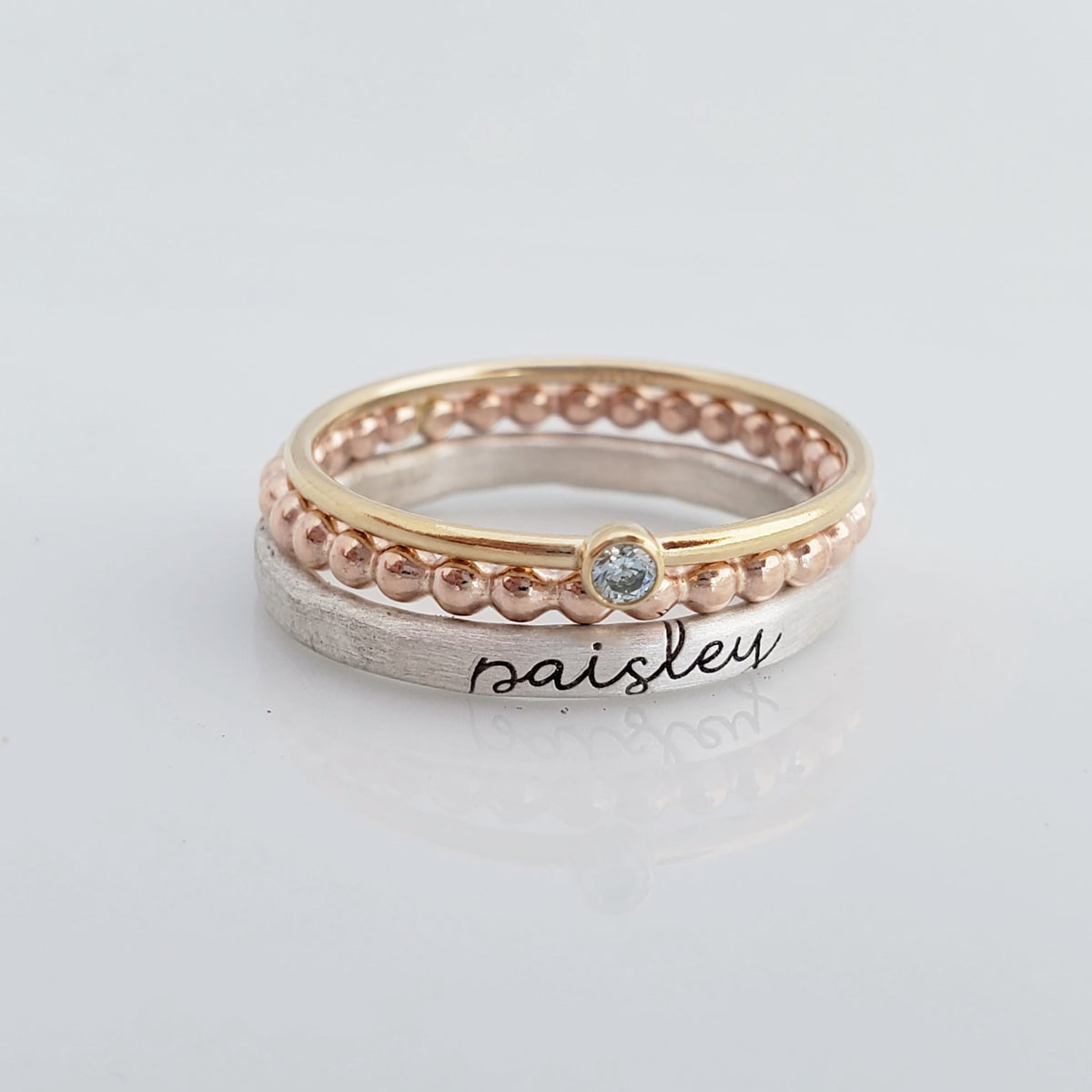 The set includes a silver ring with a brushed texture and engraved in cursive, a delicate rose gold beaded ring, and a thin yellow gold ring with a small bezel-set blue gemstone. The rings are arranged in an overlapping manner, creating a visually appealing composition with soft reflections on the surface below.
