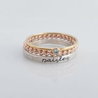 The set includes a silver ring with a brushed texture and engraved in cursive, a delicate rose gold beaded ring, and a thin yellow gold ring with a small bezel-set blue gemstone. The rings are arranged in an overlapping manner, creating a visually appealing composition with soft reflections on the surface below.