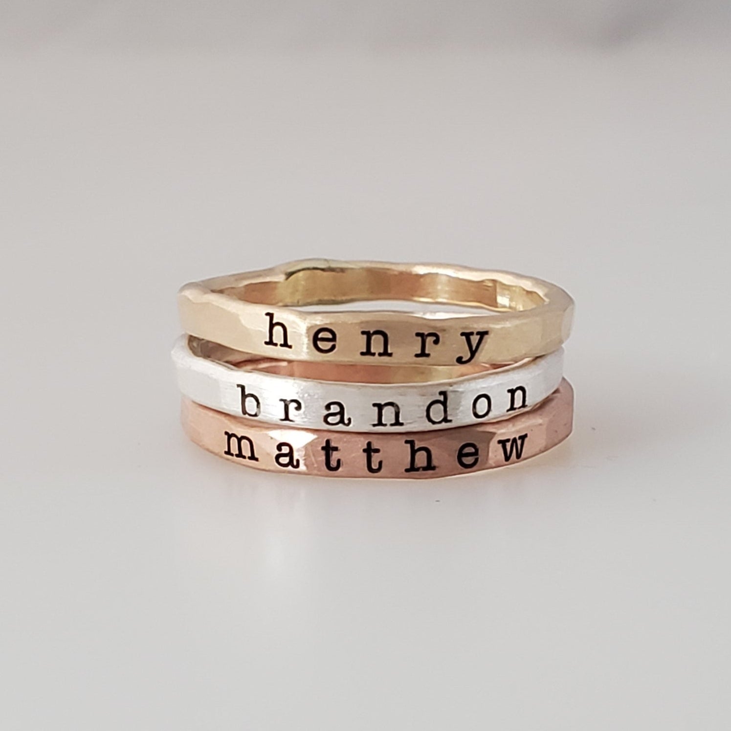 Three stacked rings made of different metals, with the names 'henry', 'brandon', and 'matthew' engraved on them, displayed on a white surface.