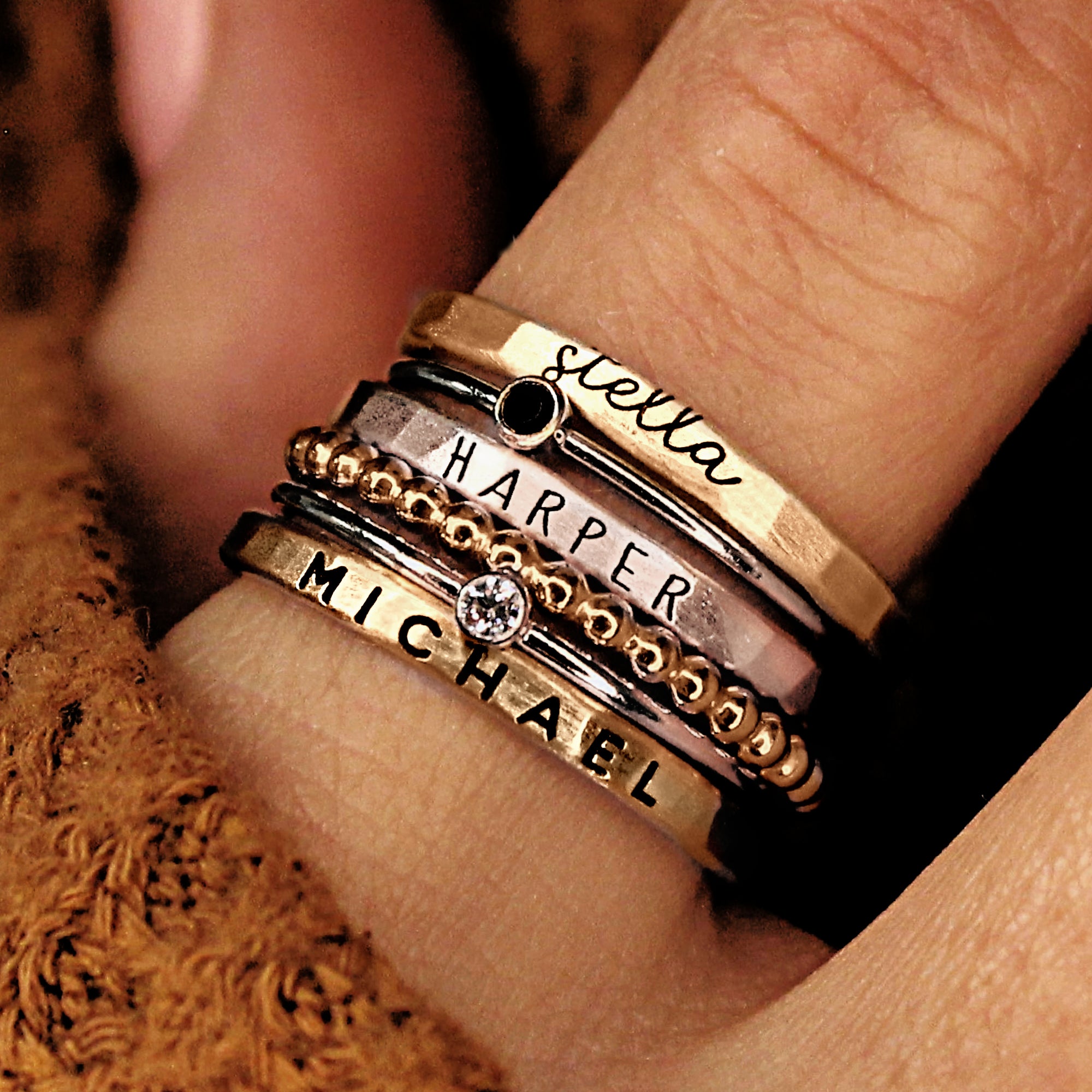 A hand wearing multiple gold-colored stacking rings with different designs, including personalized names.