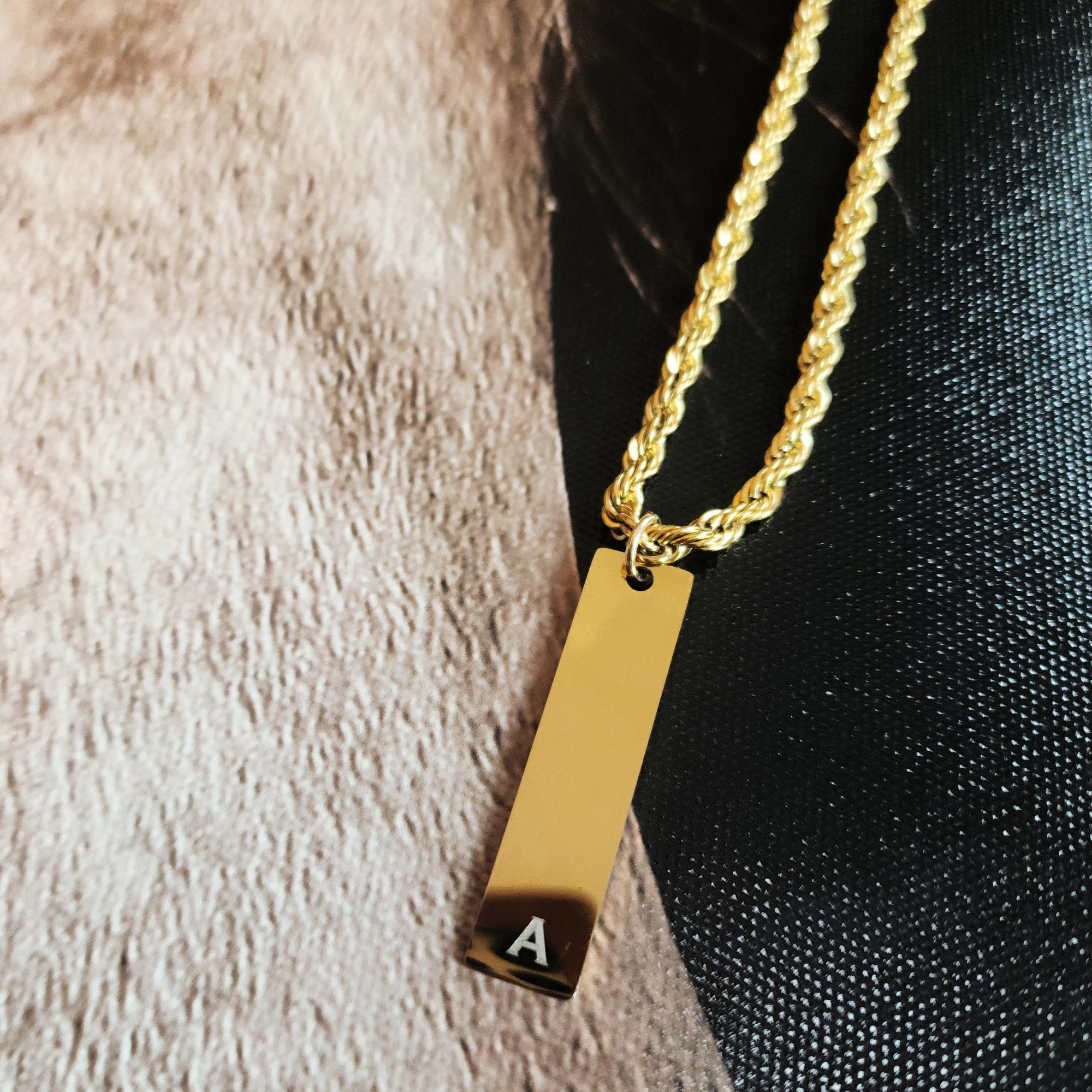 Gold necklace with a bar pendant featuring an 'A' on a textured surface.