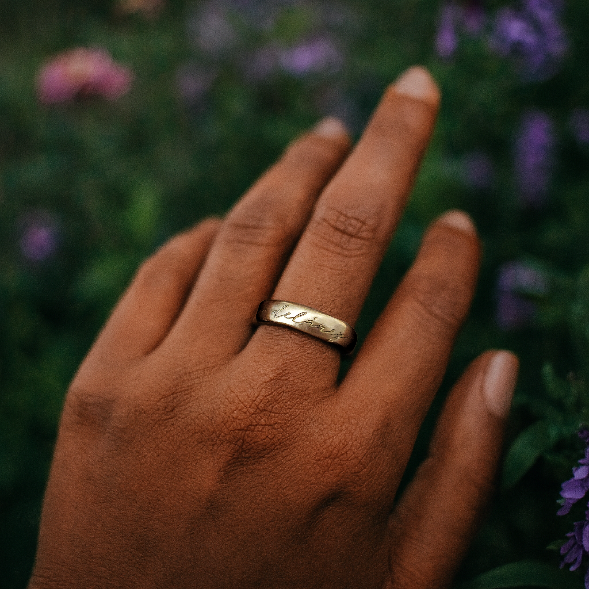 Hand wearing a gold ring with engraved text, surrounded by blurred purple flowers.