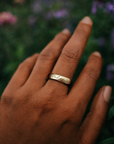 Hand wearing a gold ring with engraved text, surrounded by blurred purple flowers.