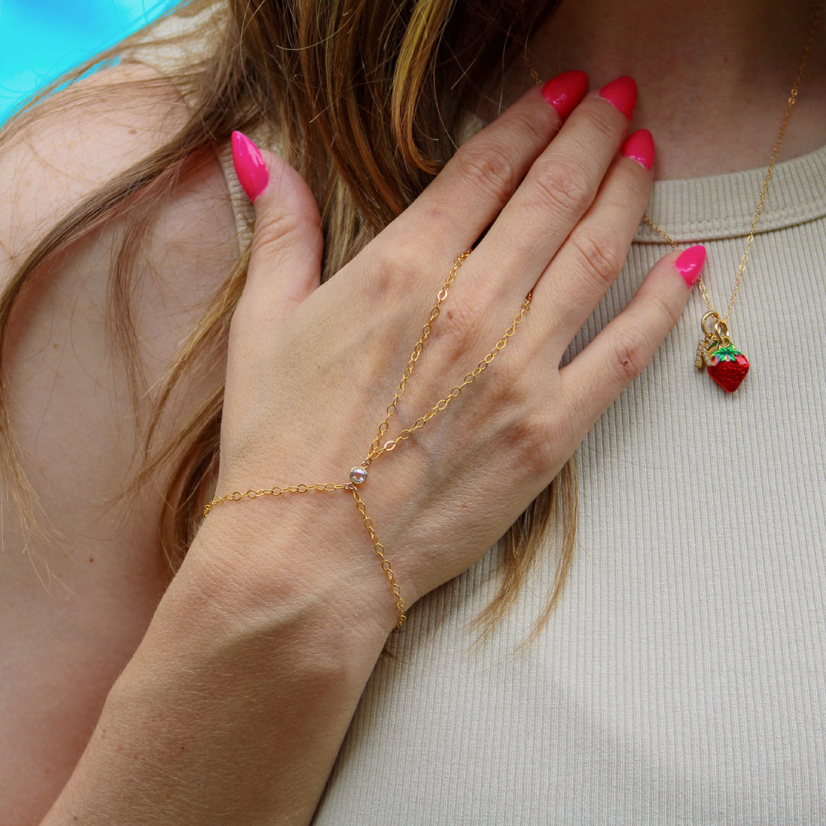 Close-up of a hand wearing 14k gold-filled hand chain and red pendant necklace, against a neutral background.