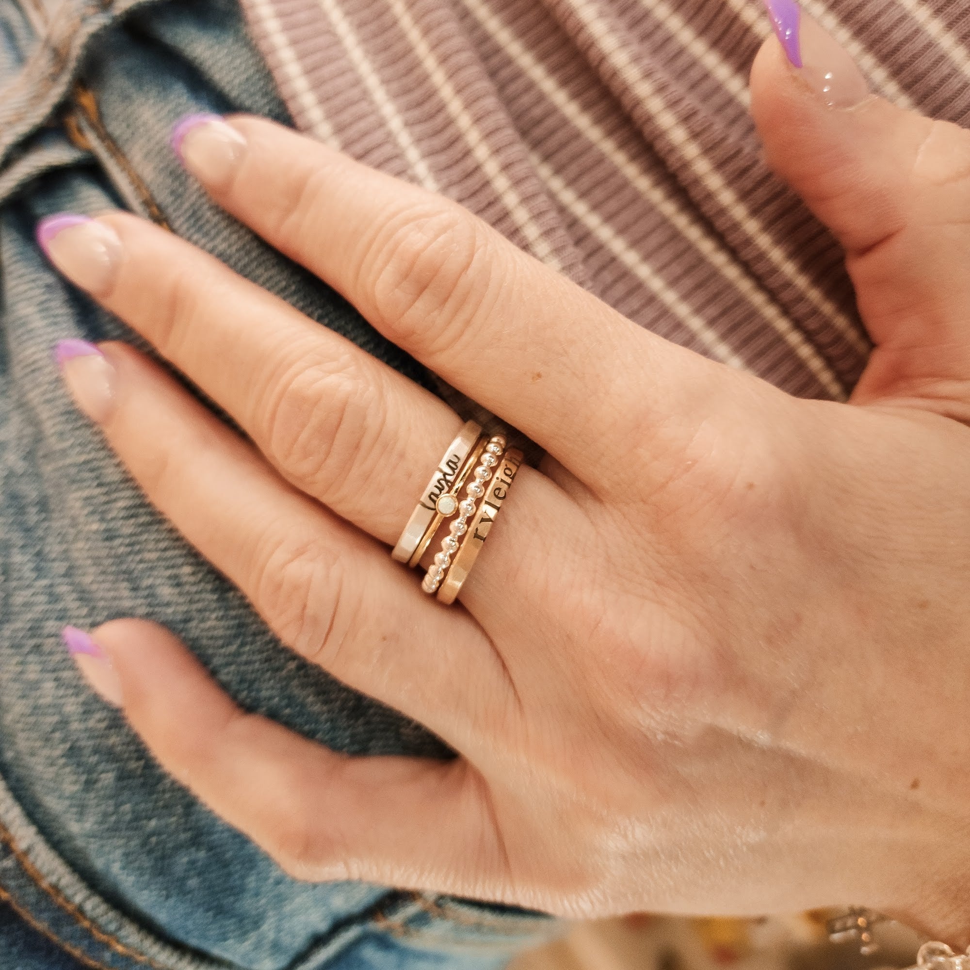 A set of four stackable rings on a white surface, including a sterling silver beaded ring, a fine silver name ring, and two 14K gold-filled rings, one with a birthstone and one with a name engraved.