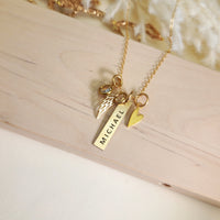 Overhead view of gold memorial necklace with “Michael” engraved bar, heart charm, angel wing charm, and birthstone on wood.