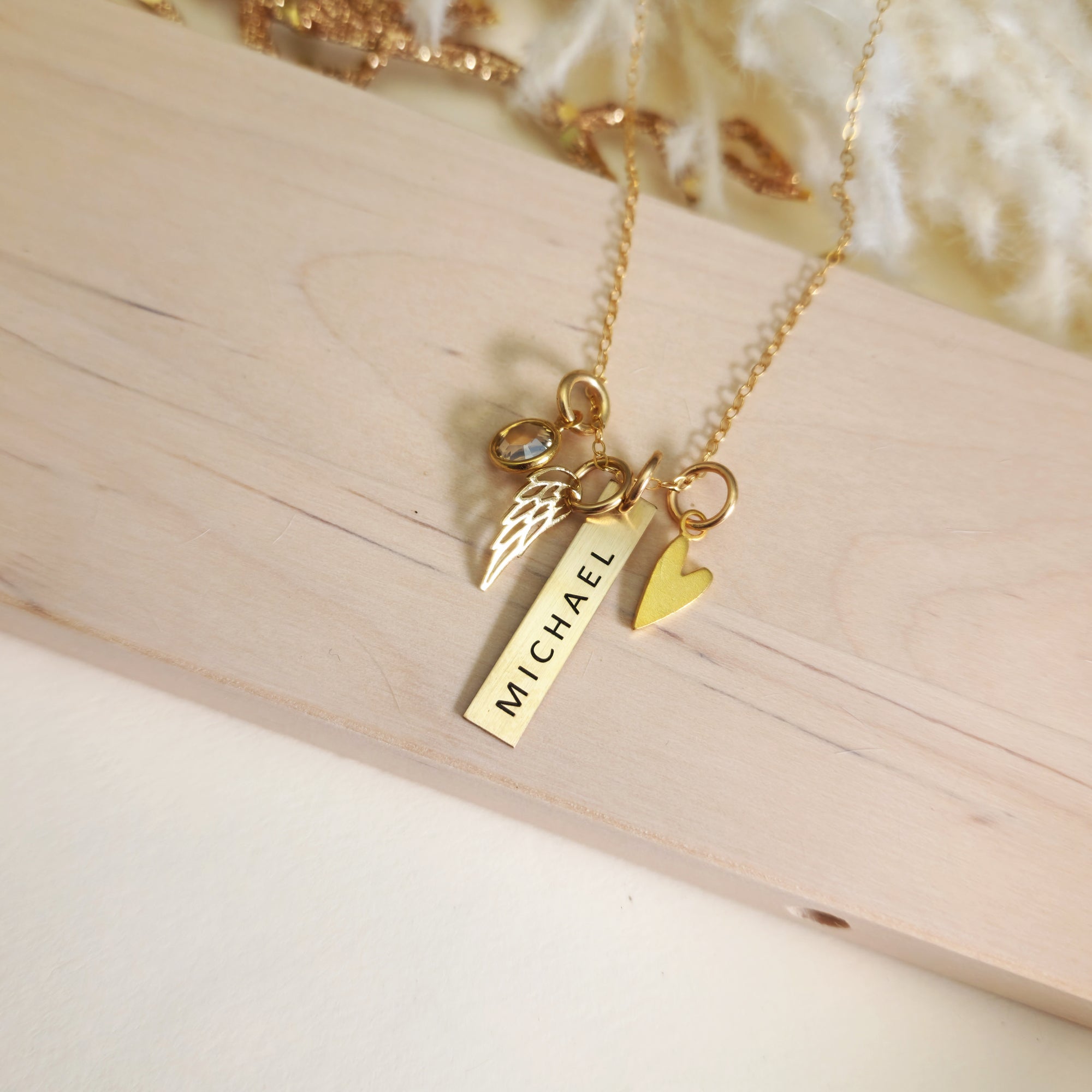 Overhead view of gold memorial necklace with “Michael” engraved bar, heart charm, angel wing charm, and birthstone on wood.