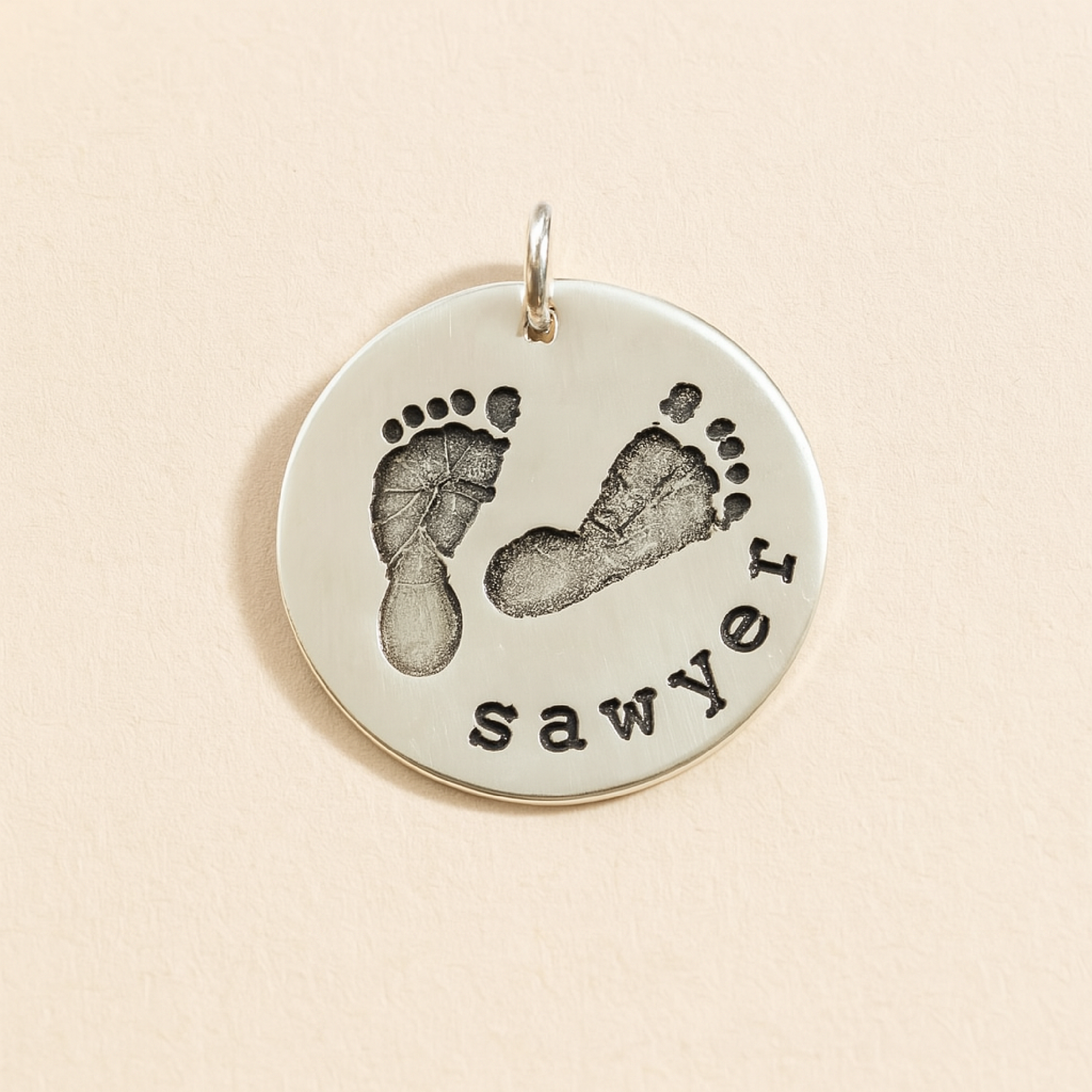 Silver pendant with footprints and the name 'sawyer' on a beige background