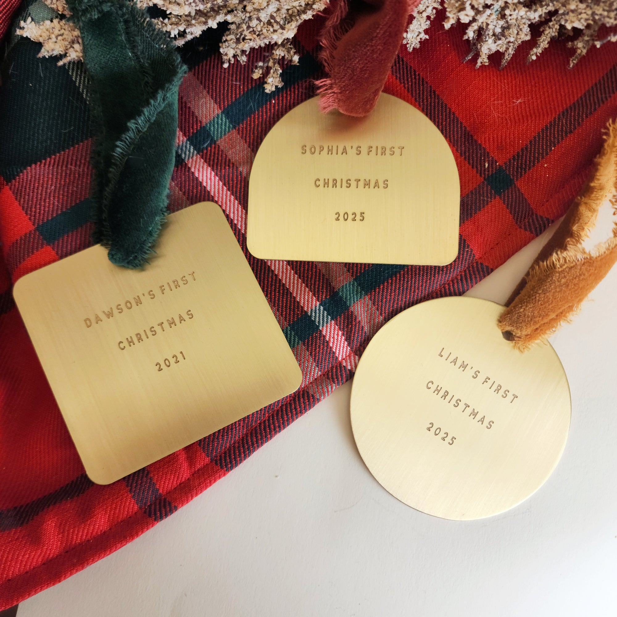 Gold 'First Christmas' tags for Sophia, Dawson, and Liam on a red plaid fabric background.