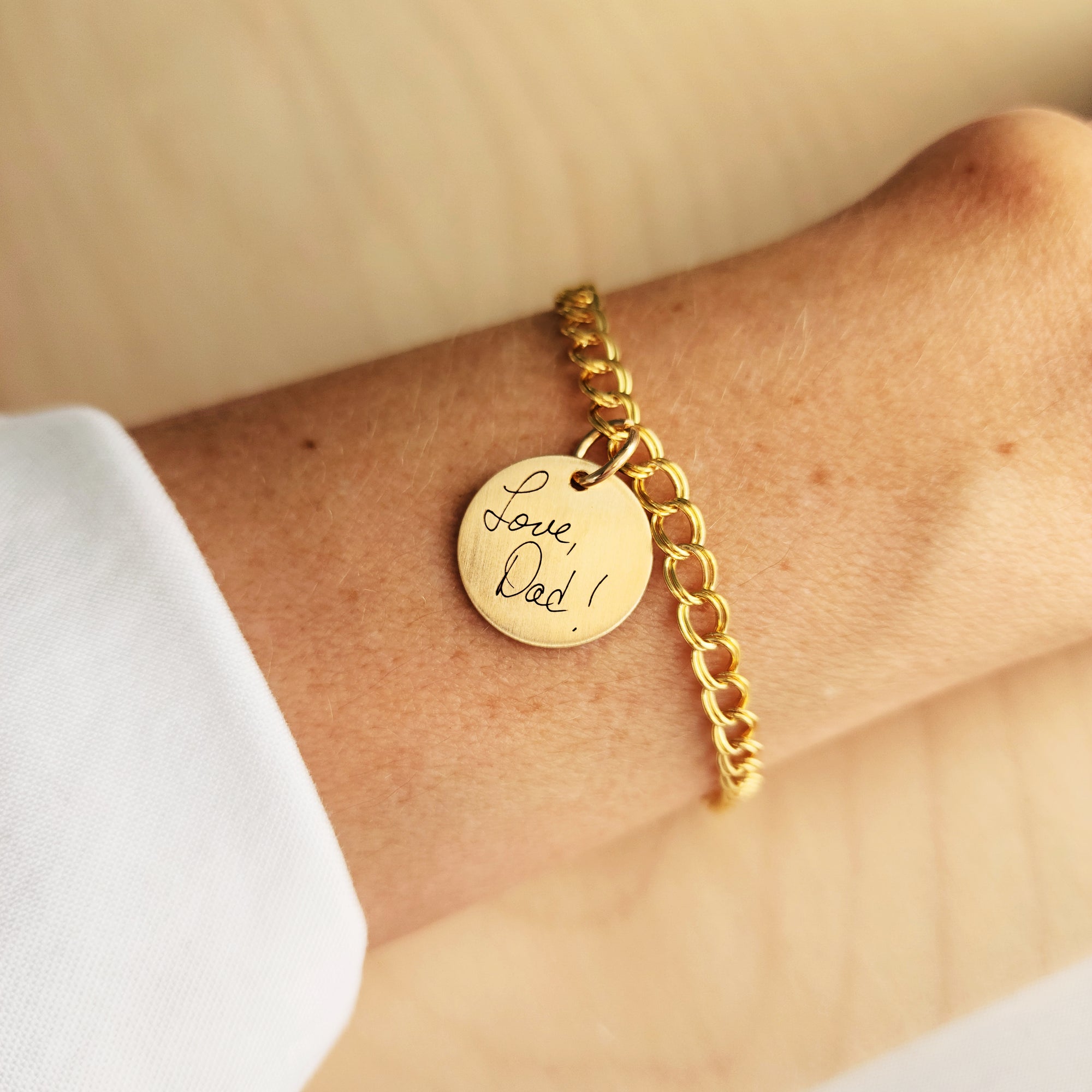 A gold charm bracelet with a round pendant engraved with 'Love Dad' on a person's wrist, featuring a classic gold chain.