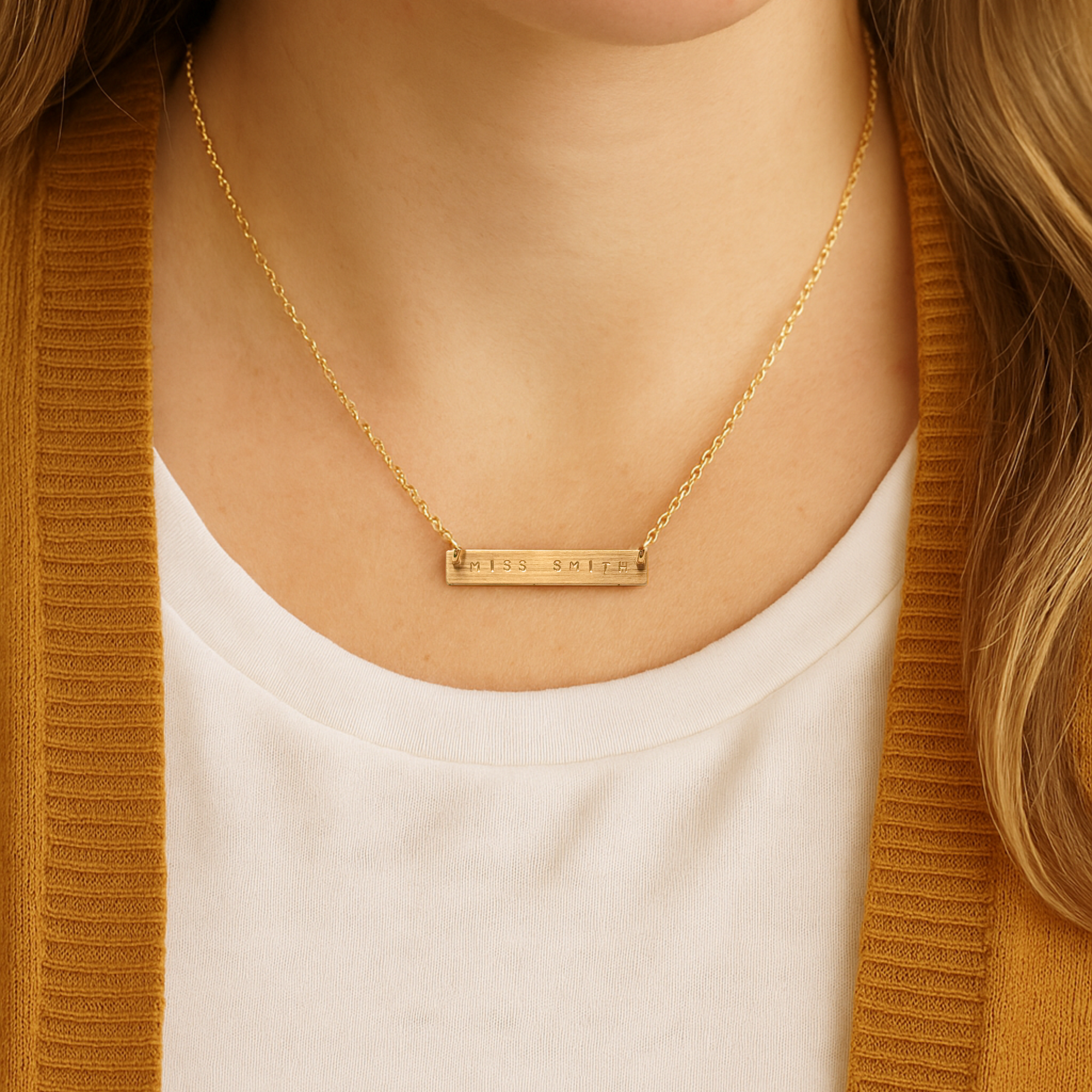 Woman wearing a personalized gold bar teacher necklace engraved with MISS SMITH on a cable chain, styled with a mustard cardigan and white shirt
