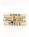 A set of three stackable rings made of yellow gold, with two gemstones, and personalized engravings including 'AVON', 'class of 2022', and 'MAREN'.