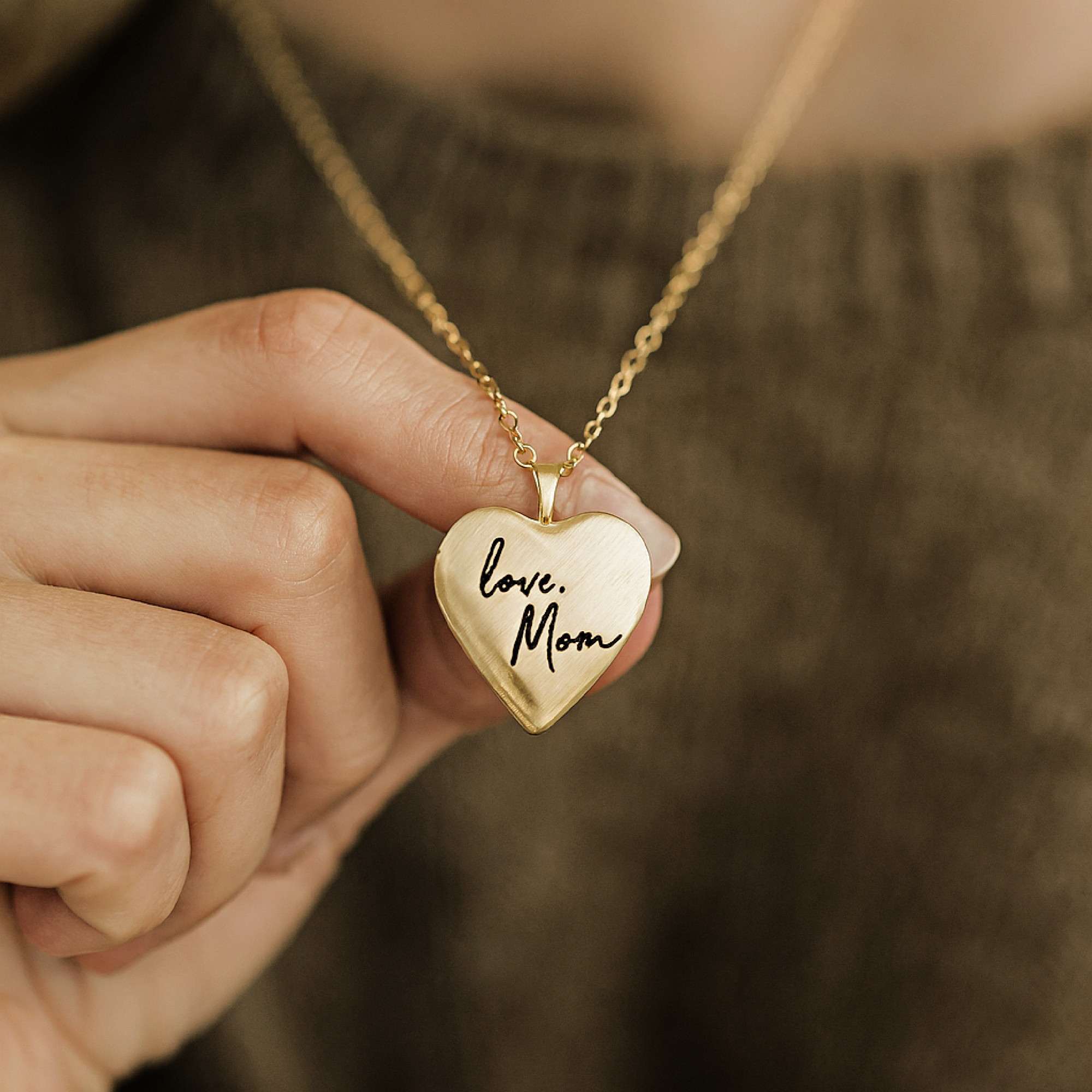 Heart-shaped gold necklace with 'love, Mom' engraving held by a hand against a blurred background