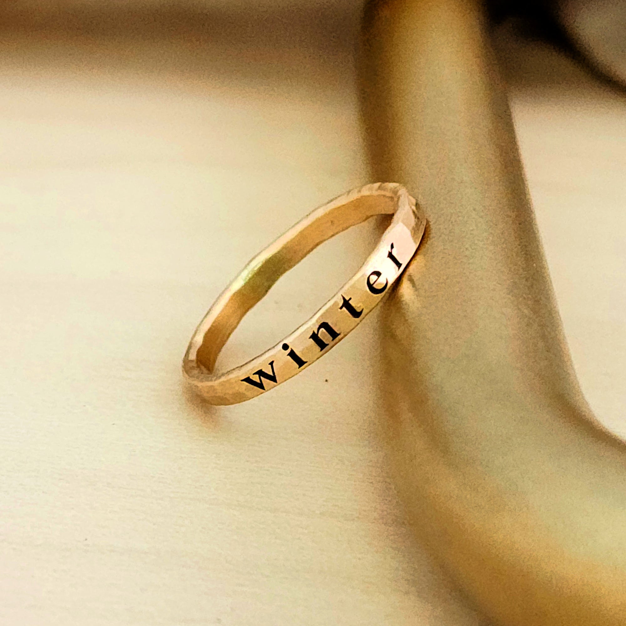 A 2mm gold stacking name ring is resting on a gold bar.  The ring has a name engraved in dark font on the center of the ring.  There is a visible yellow solder spot on the back of the ring.