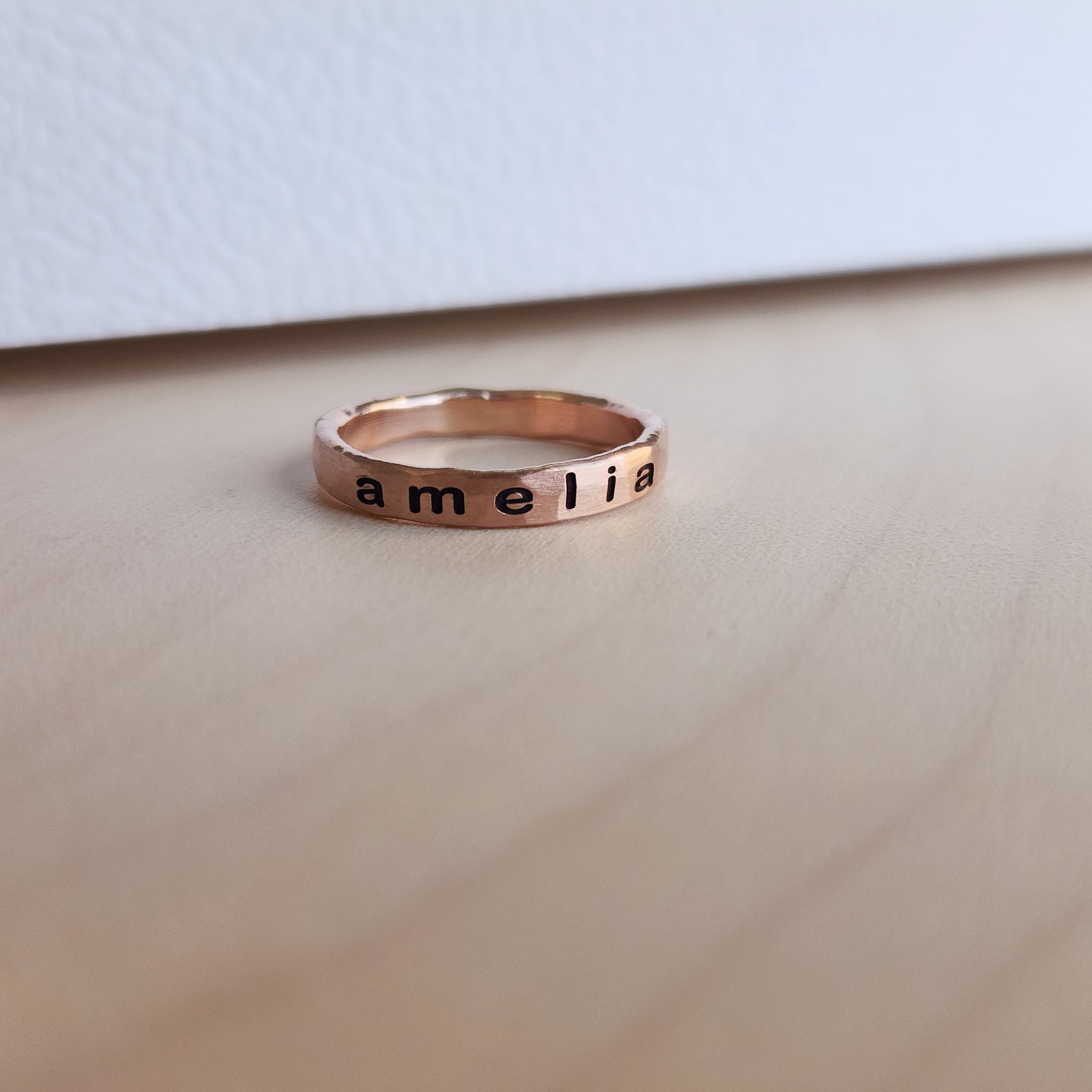 A rose gold filled personalized stacking ring with the name 'amelia' engraved on it, displayed on a wooden surface.