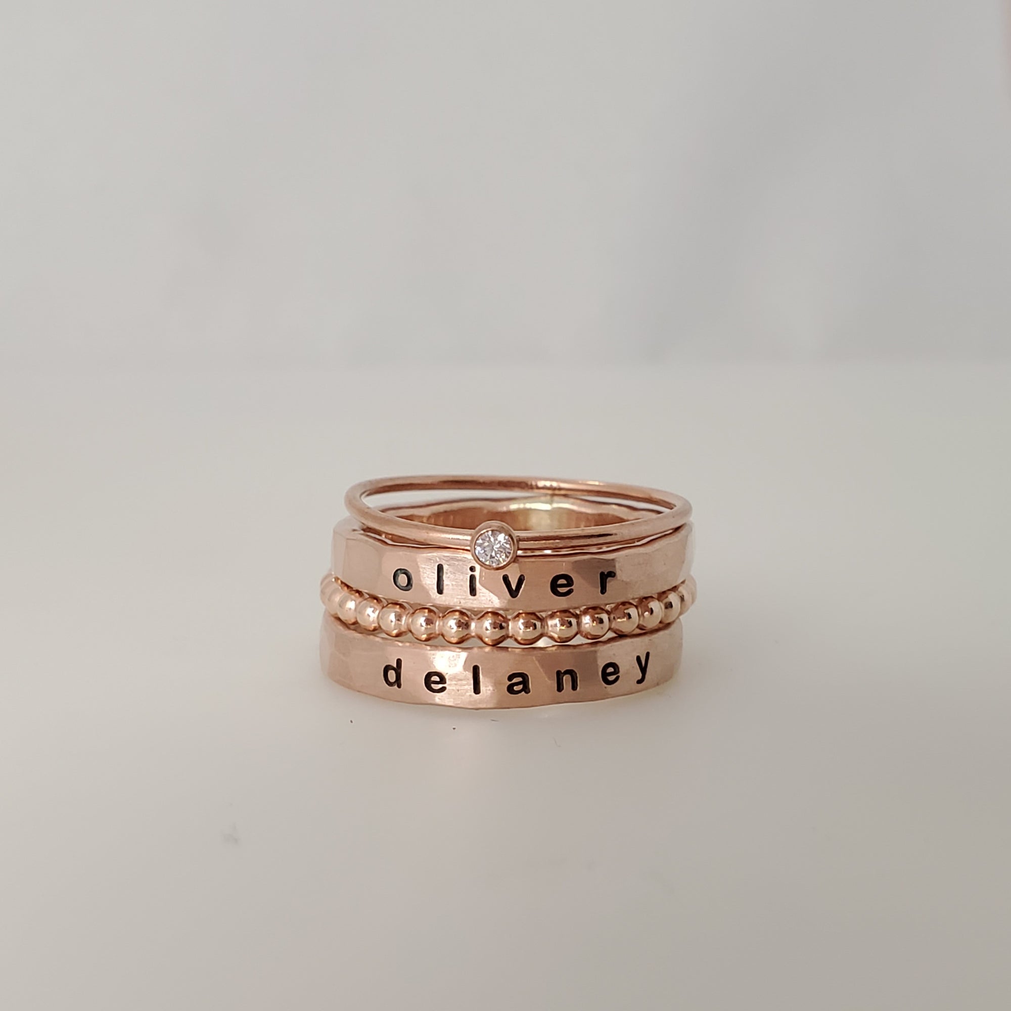 A set of stacked silver rings, with one ring engraved with the name 'OLIVER' and another with 'DELANEY'. The set includes a ring with a small diamond at the top, and a beaded spacer ring.