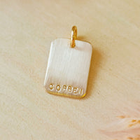 Personalized rectangle charm stamped with the name “CORBEN,” shown without a chain, on a warm-toned wooden background with dried flowers.