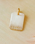 Personalized rectangle charm stamped with the name “CORBEN,” shown without a chain, on a warm-toned wooden background with dried flowers.