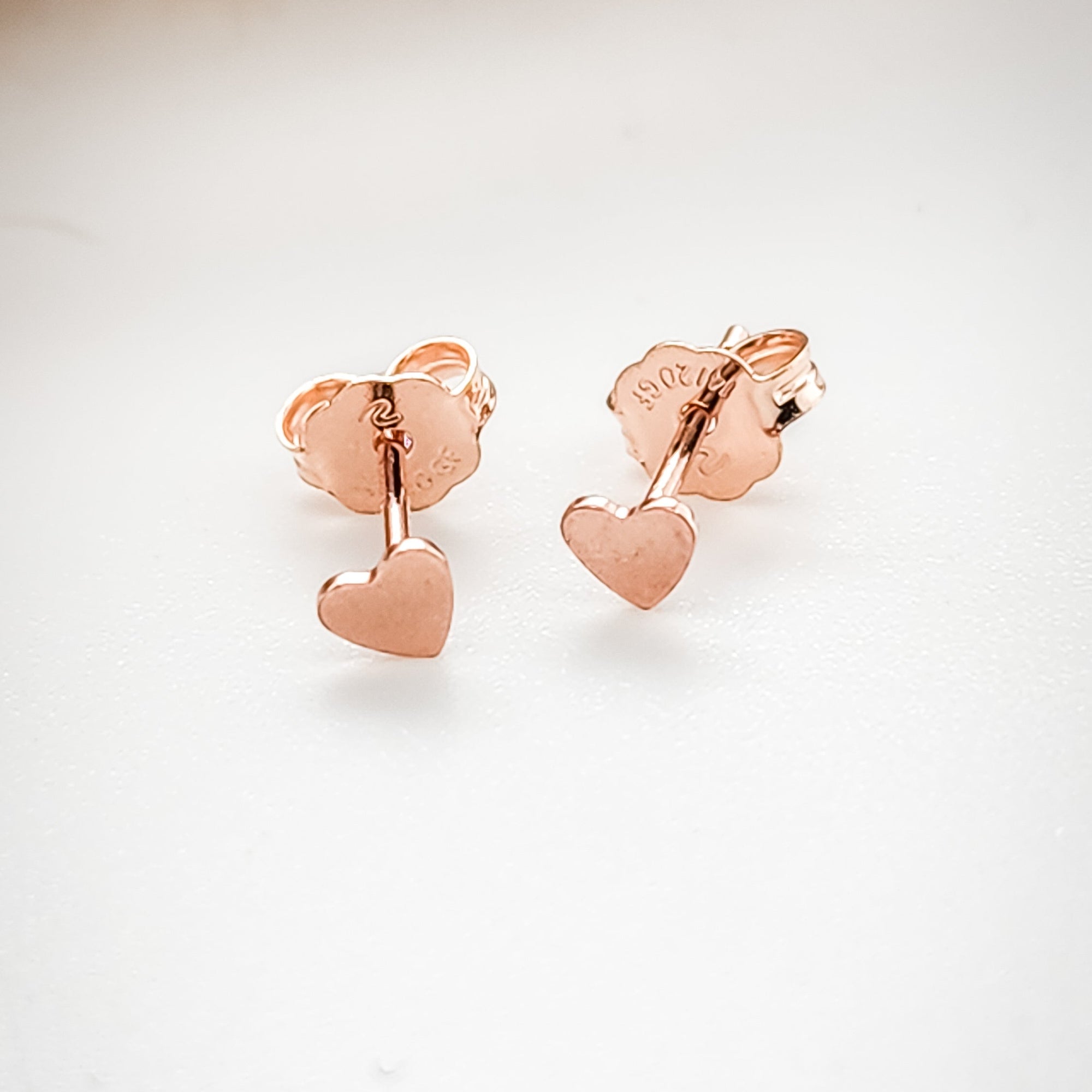 A pair of rose gold heart stud earrings on a white background.