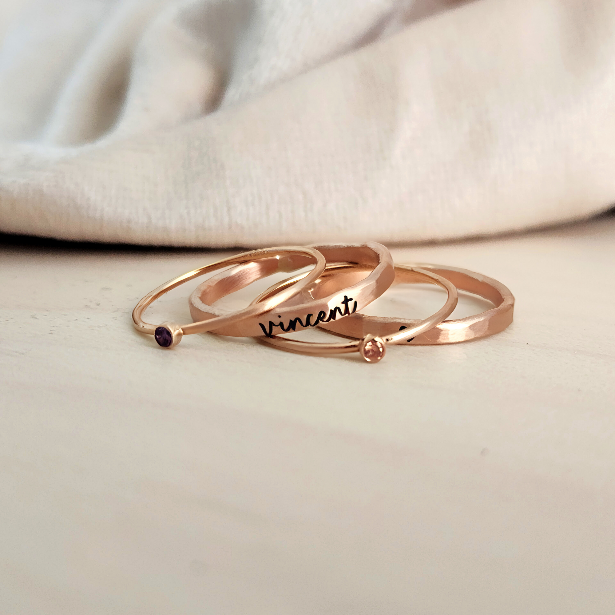 A set of three gold-colored rings, two with engraved names and one with a birthstone, displayed on a person&#39;s finger.