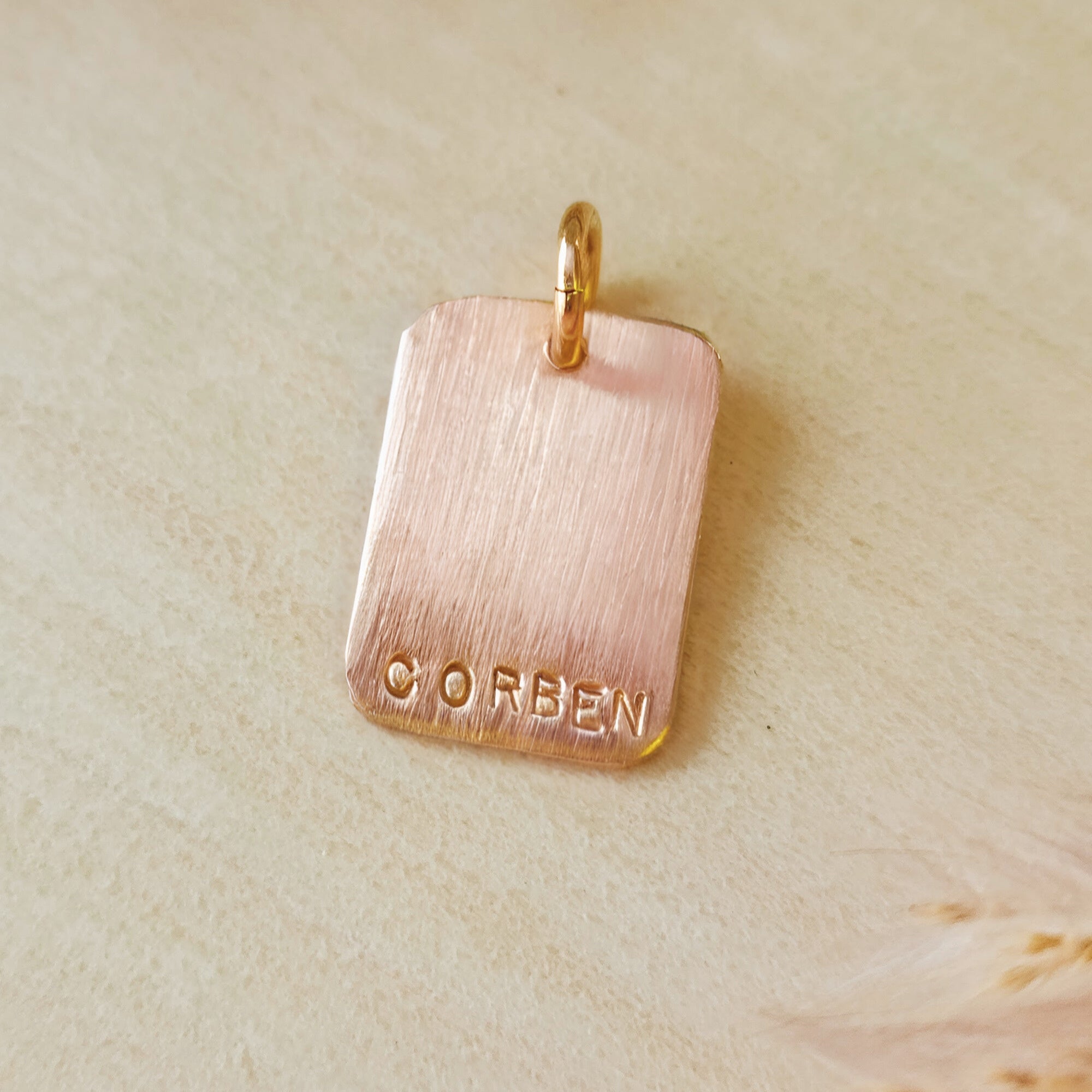 Rose gold pendant with 'CORBEN' engraving on a beige background