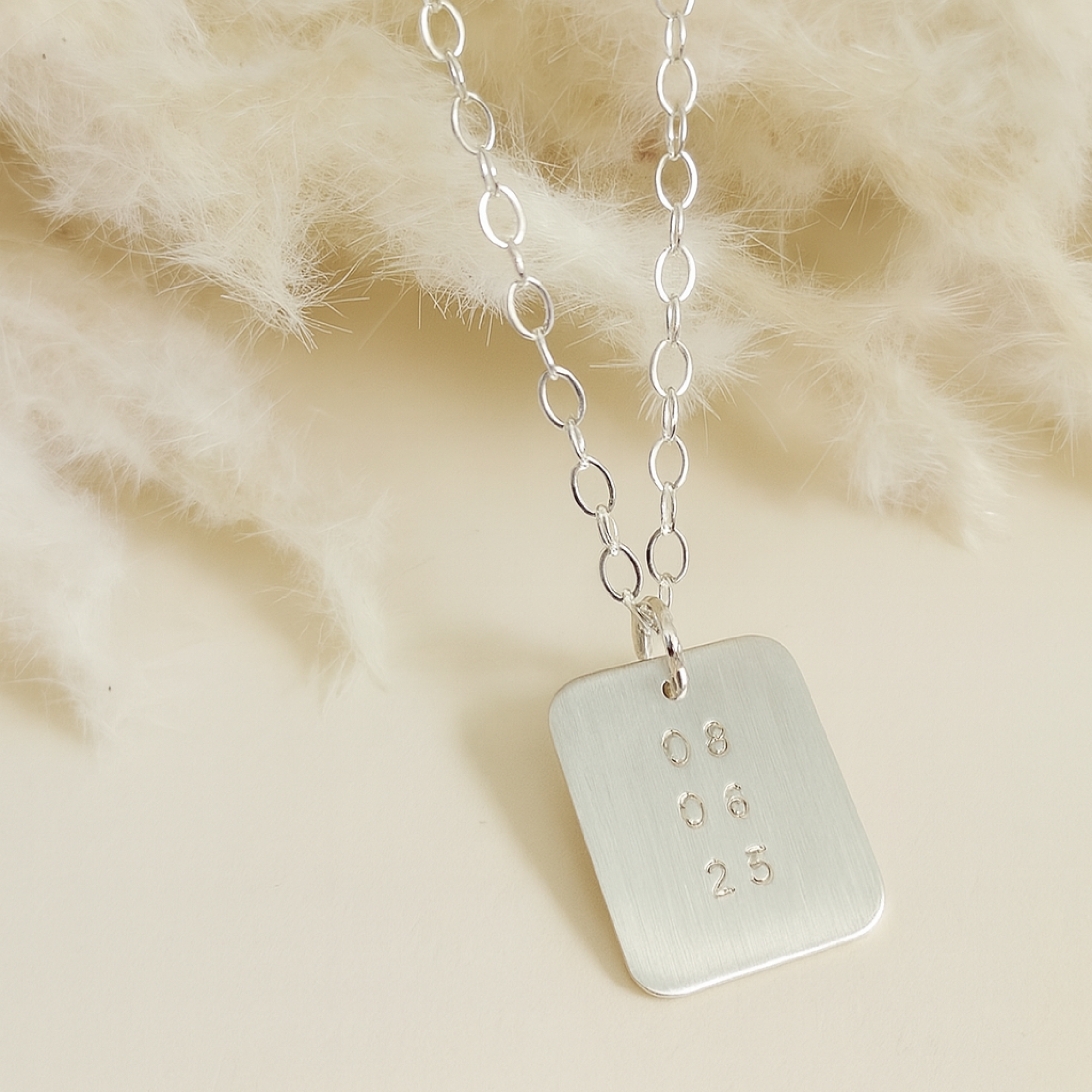 Silver date-stamped pendant necklace displayed on pampas grass background