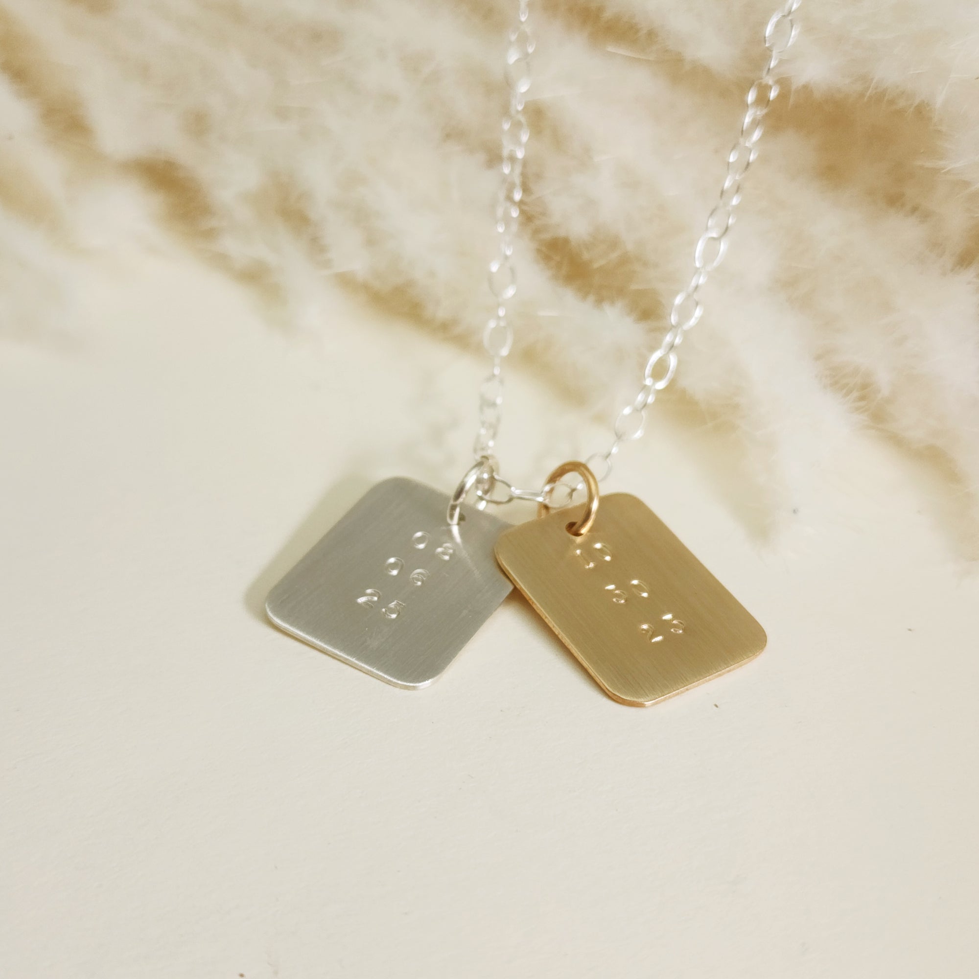 Silver and gold date-stamped pendant necklaces layered together on a silver chain, displayed on a cream background.