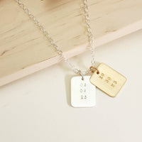 Two rectangular date-stamped pendants, one gold and one silver, on a silver chain displayed on a light wood background.
