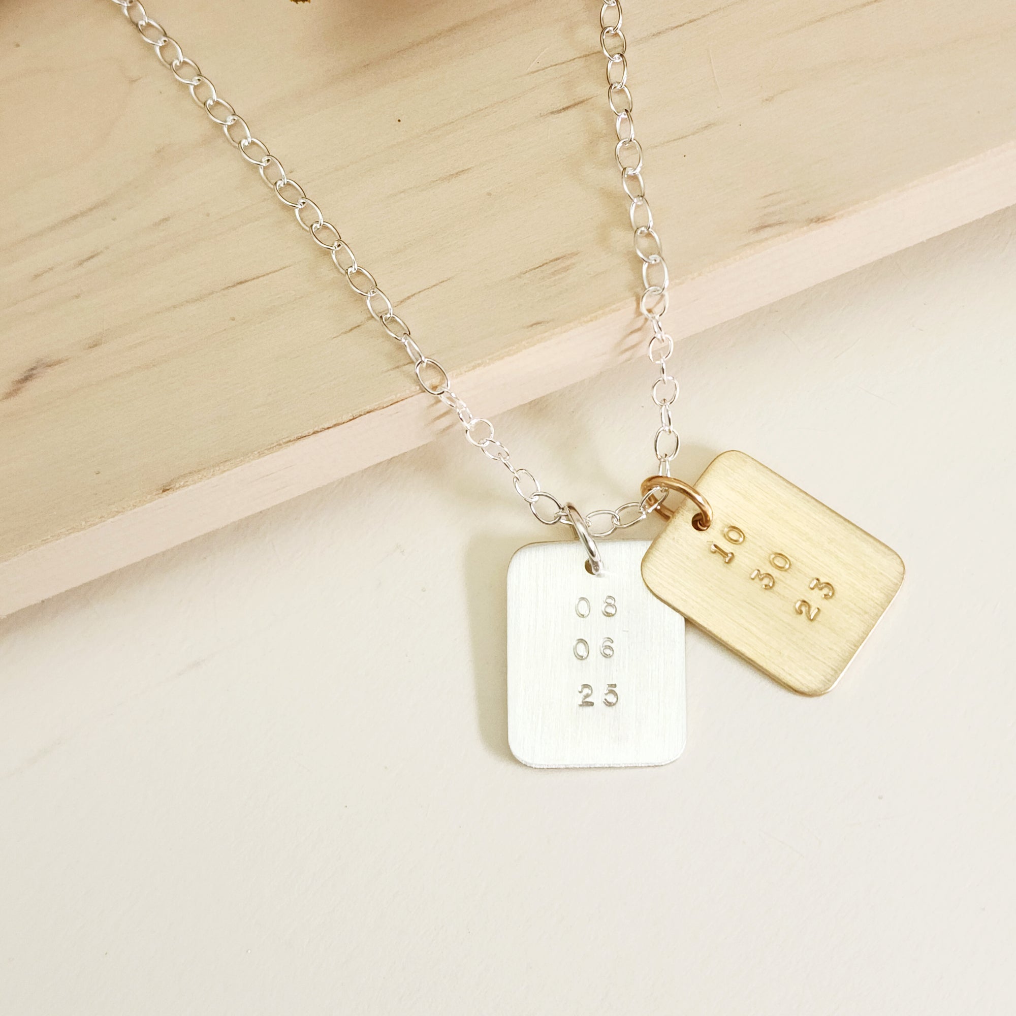 Two rectangular date-stamped pendants, one gold and one silver, on a silver chain displayed on a light wood background.