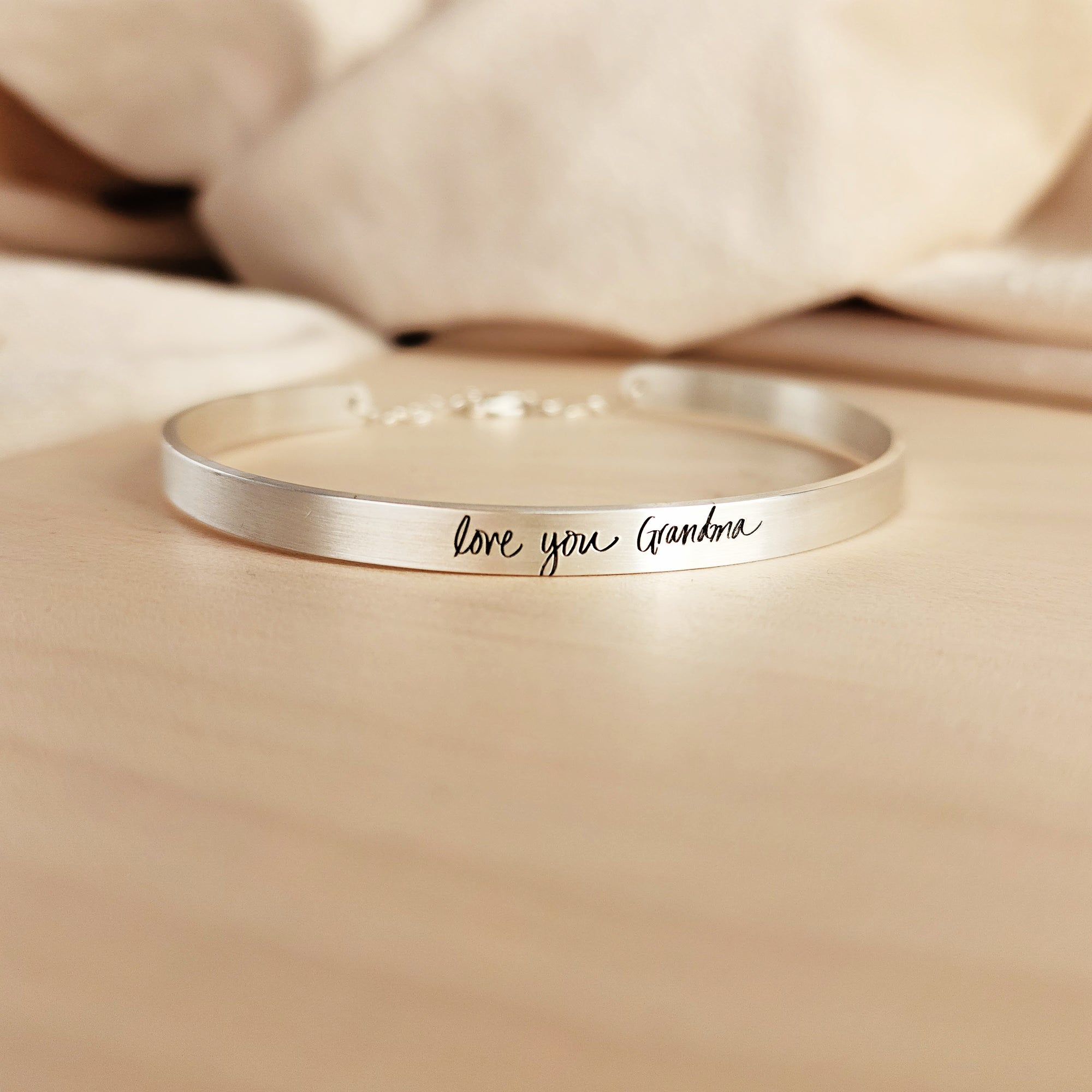 A silver cuff bracelet with custom handwritten engraving, displayed on a wooden surface.