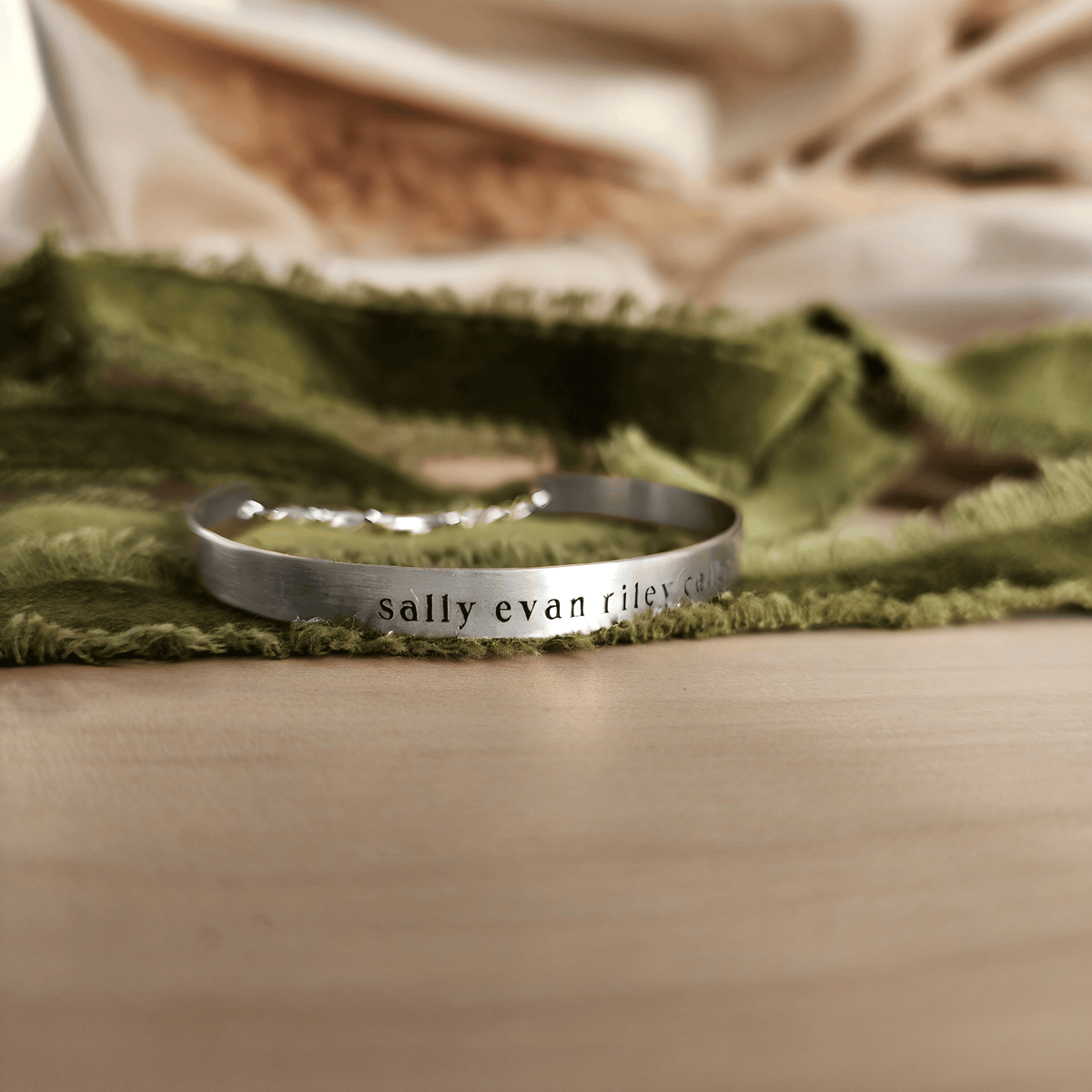 A gold-colored cuff bracelet with custom names engraved on it, displayed on a white surface with a green textured object to the side.