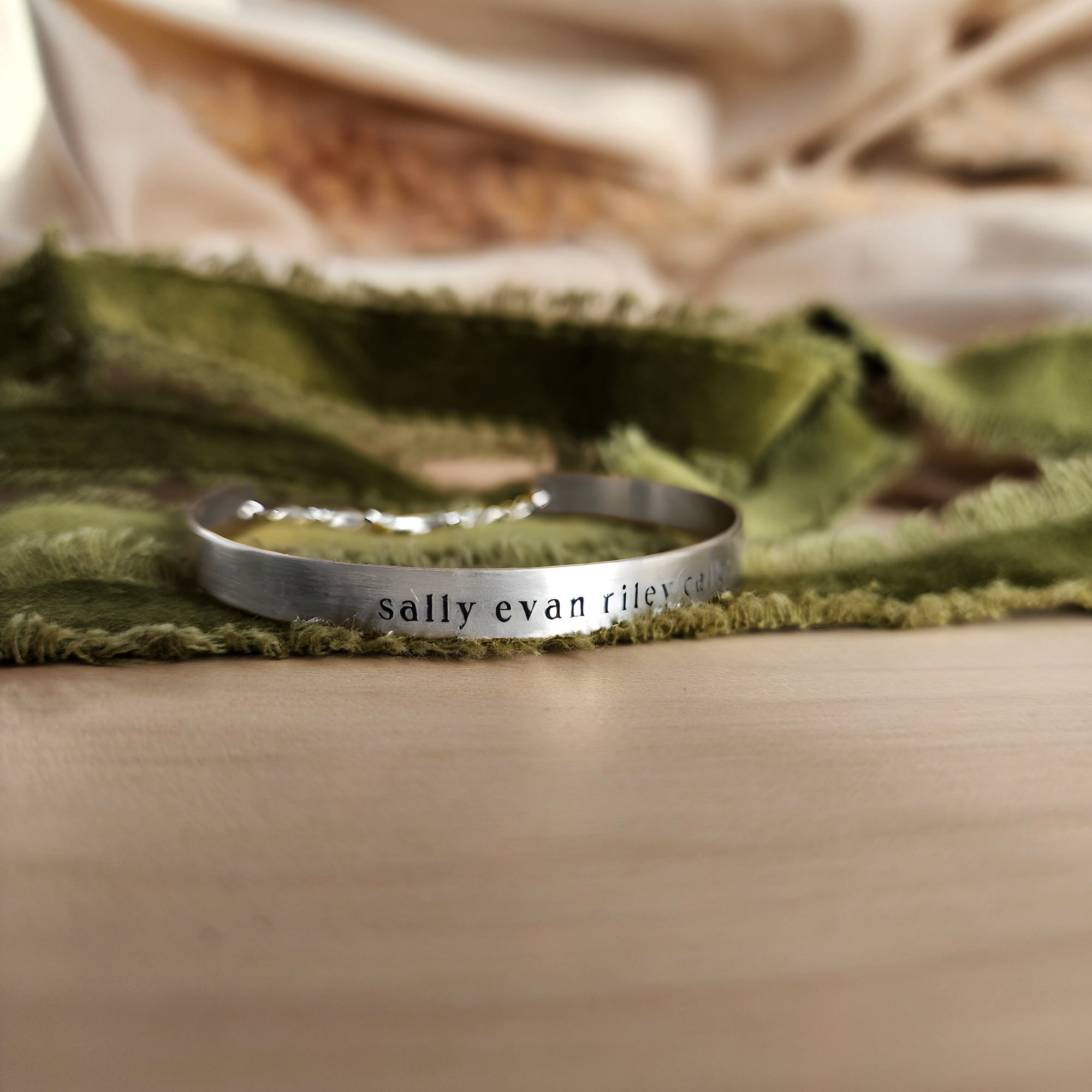 A gold-colored cuff bracelet with custom names engraved on it, displayed on a white surface with a green textured object to the side.