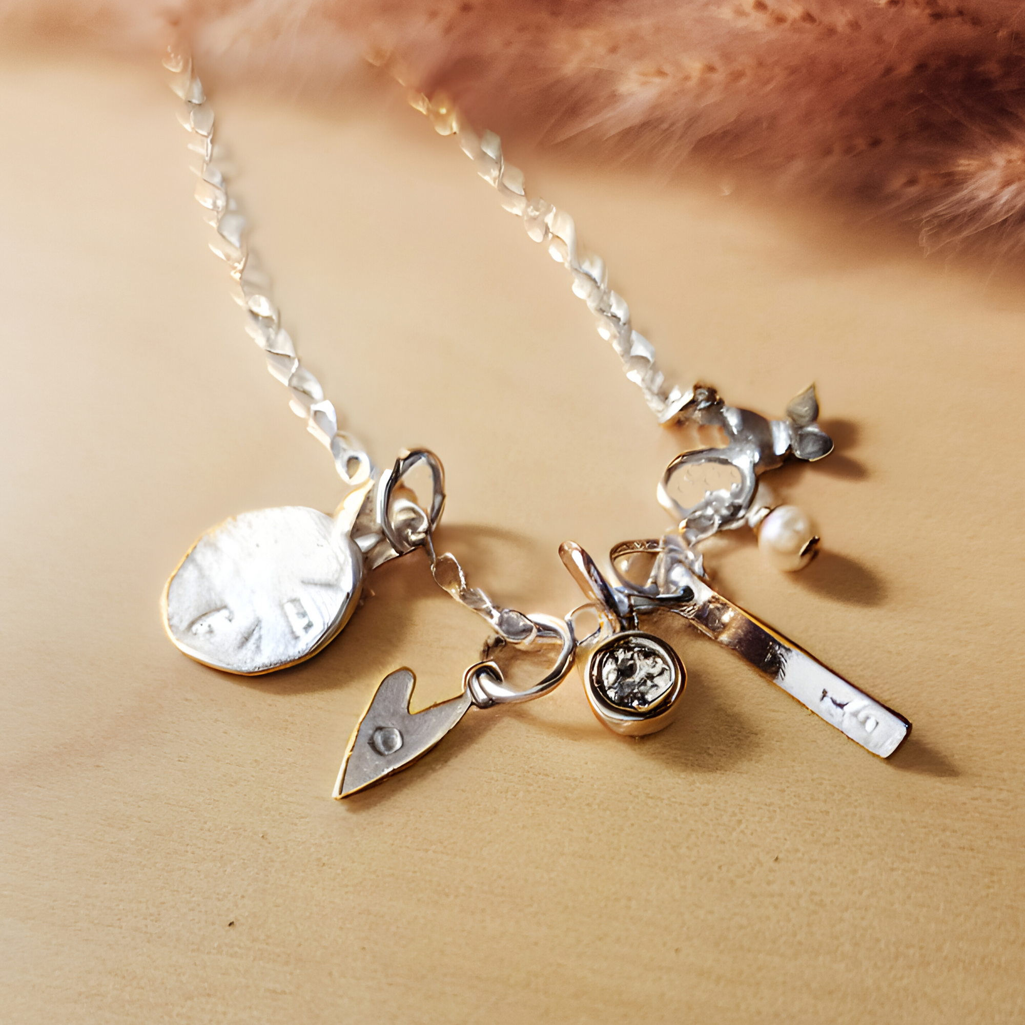 Close-up of a silver charm necklace displayed on a light wood surface with soft pink pampas grass in the background. The necklace features multiple silver charms, including: a round disc stamped with "GAB", a small heart with an engraved letter, a bezel-set clear crystal charm, a vertical bar engraved with "10", a small pearl dangle, and a delicate leaf charm. The chain is a shiny flat curb style, giving the piece a polished, customized look.