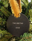 Round black ornament with 'The Smiths' and '2025' text, hanging on a Christmas tree.