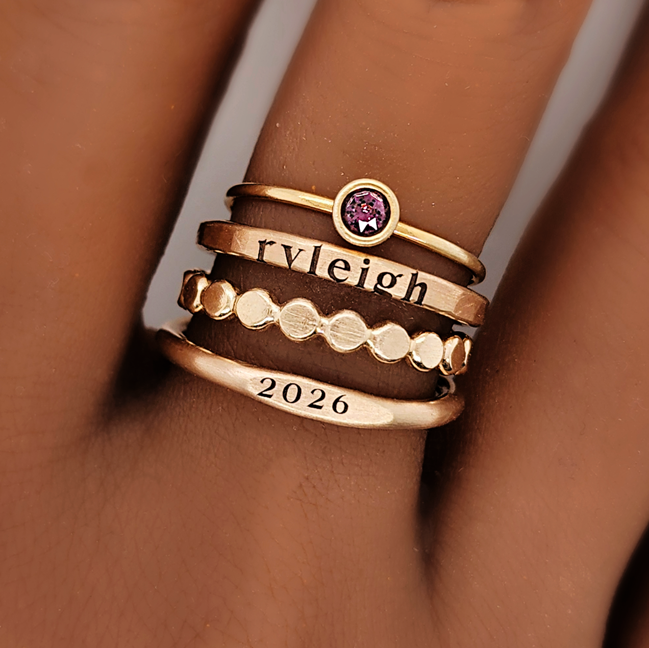 A set of stackable gold rings on a person's hand, featuring personalized engravings such as a name and the year 2026.