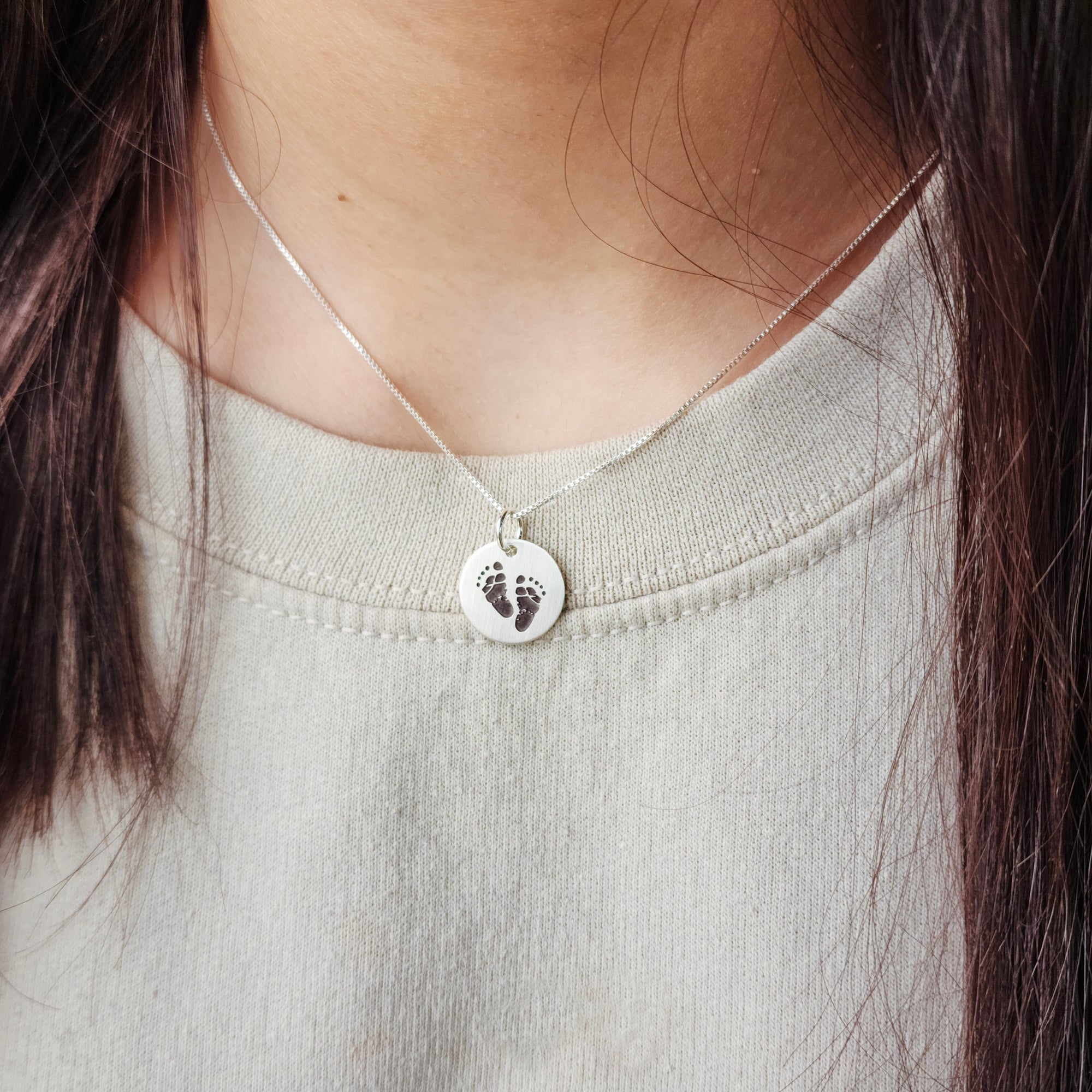 A person wearing a grey sweater and a necklace with an engraved footprint pendant on a silver chain.