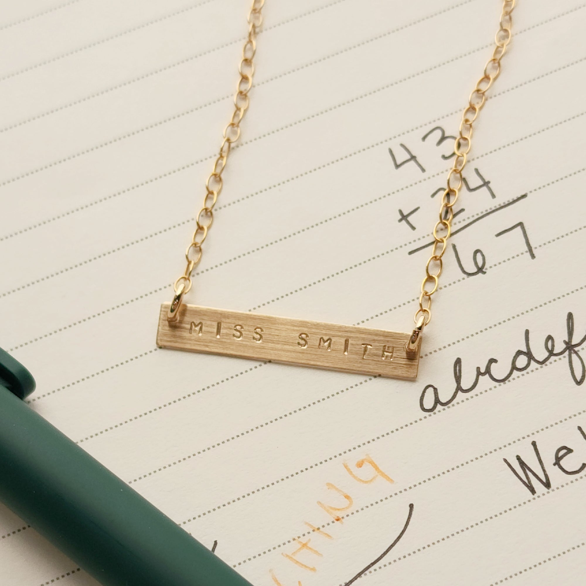 Personalized gold bar teacher necklace stamped with MISS SMITH, photographed on lined school paper with handwritten math and alphabet.