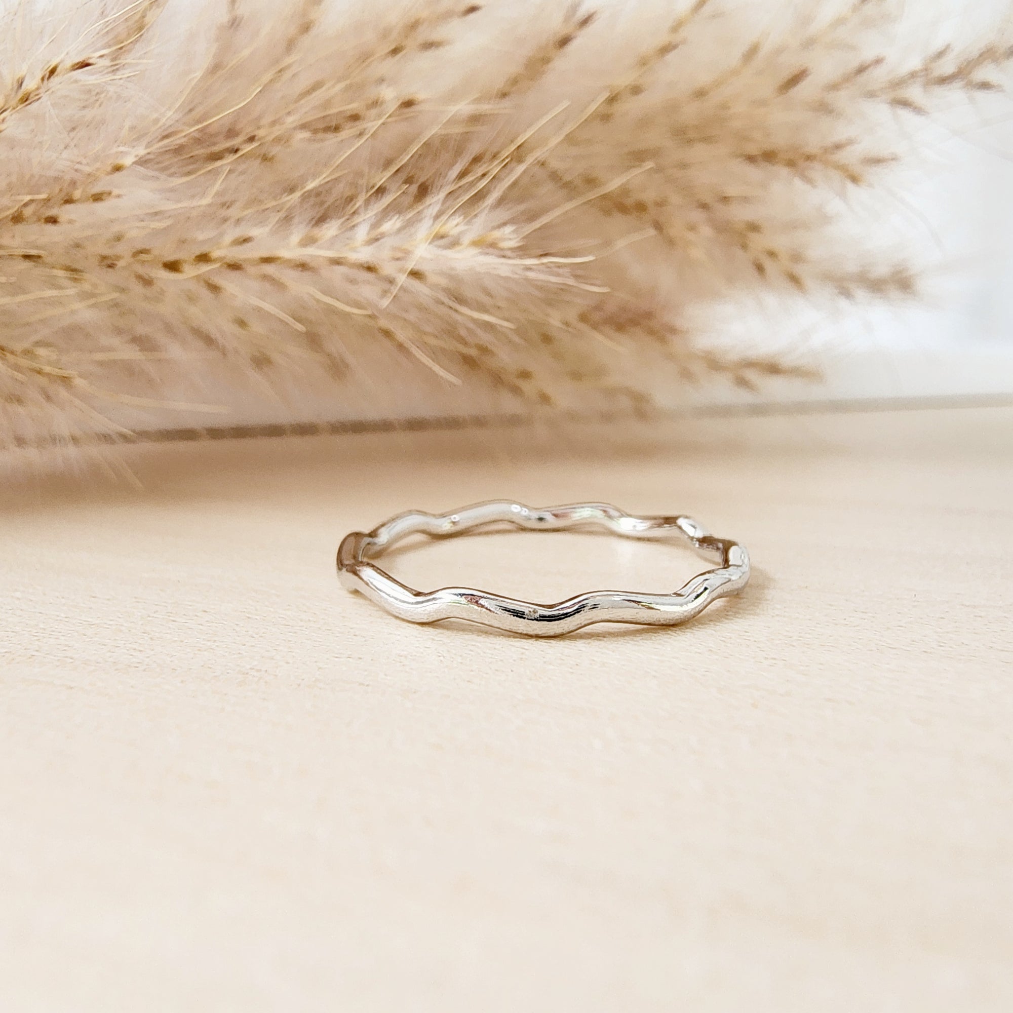 A silver stacking ring with a wavy design, displayed against a neutral background with dry plants in the proximity.
