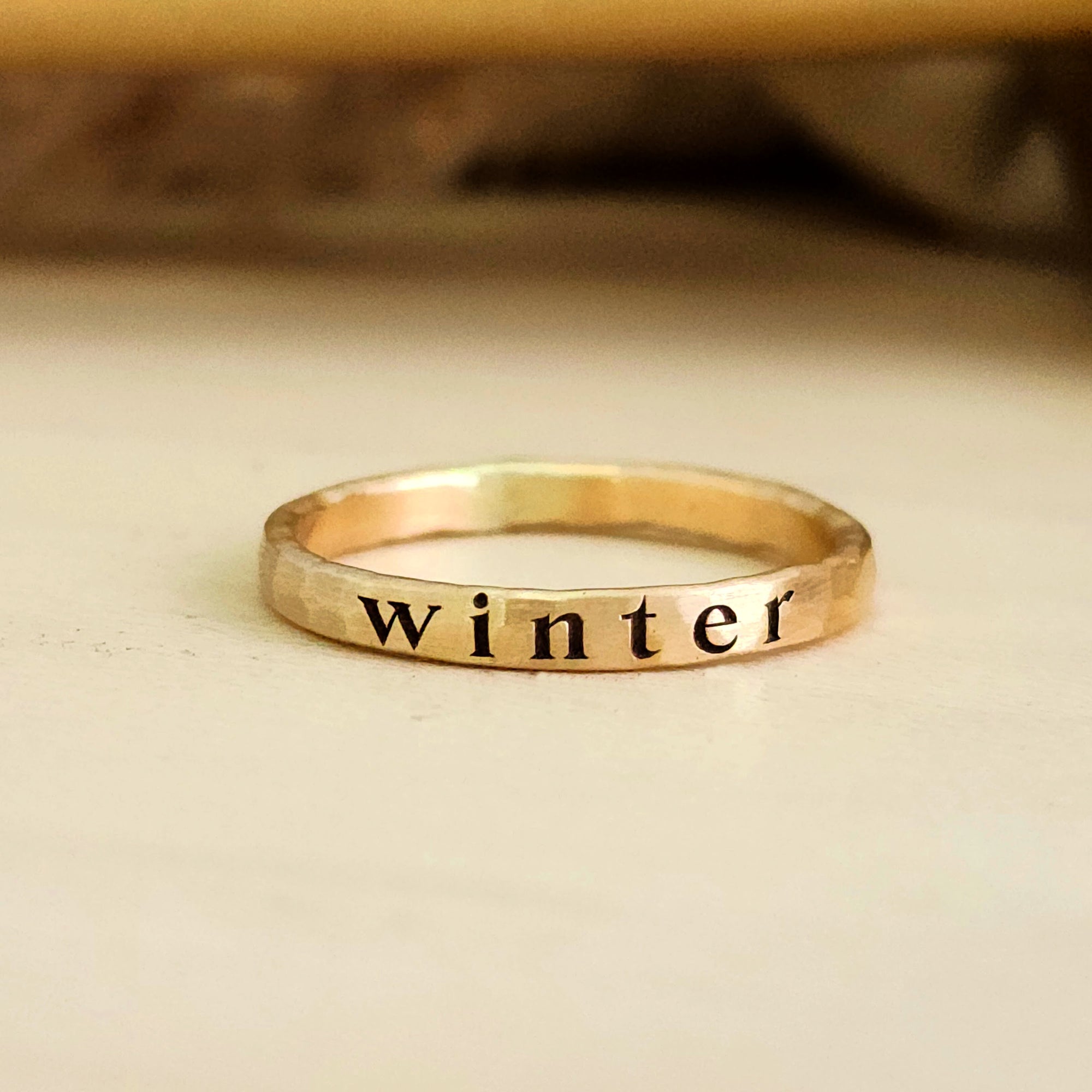 yellow gold ring sitting on a light wood table.  Engraved deeply on the ring in serif font, the name "winter".  Shadows in background.