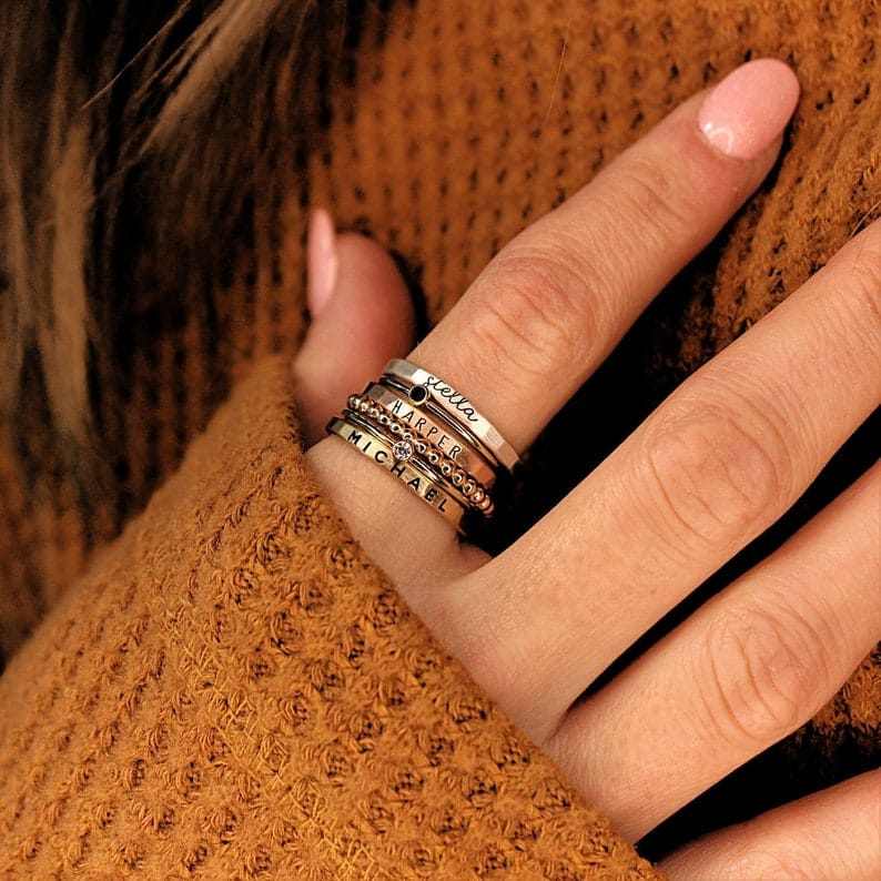 Close-up of hand wearing personalized stacking rings with names, promoting custom design your own ring set in silver and gold materials