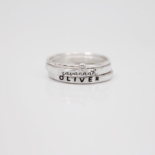 A set of three rings, including two name rings in different fonts and a clear birthstone ring, displayed against a white background.